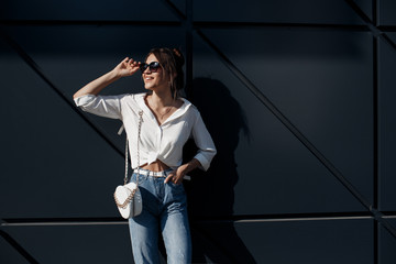 A brunette woman with long hair in fashionable in blue jeans and a white shirt posing outdoors.Fashion photo concept. Woman near business center.Beautiful female business center worker near her office