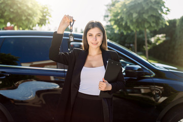 Beautiful girl with car key in hand. Caucasian woman car seller holding car keys, standing in front of new black car outdoors in vehicle trade fair. Auto rental or sales concept.