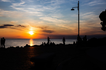 Dramatic and beautiful sunset at the coast line of the mediterranean sea in trieste Italy with silhouettes of a couple and people sitting on the beach.