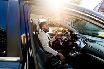 Young handsome African businessman using mobile phone, speaking through the loudspeaker, while sitting inside a luxury car