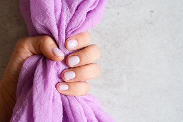 a woman's hand with pale purple painted nails holds a lilac cotton fabric on a gray concrete background. space for text