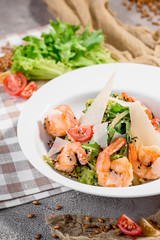 Traditional Caesar salad with shrimp, tomatoes, lettuce, egg, parmesan and croutons in white plate on a light concrete background in beautiful composition with napkins and fresh vegetables. Close up