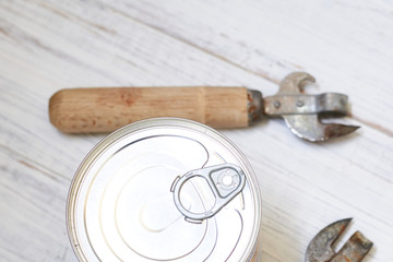 Canned food with a key for opening and an old can opener on a light wooden background.