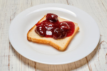 Toast with strawberry jam on a white plate on a light wooden background.