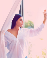 The beautiful woman wearing white shirt standing beside white curtain,looking outside window,portrait of model posing,vintage and art tone,blurry light around