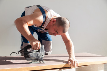 Closeup hand of carpenter, woodworker with professional cutting tool fretsaw or jigsaw, cut wooden tabletop, sawing plank, brown filings, sawdust. Concept cut hole for sink in the kitchen