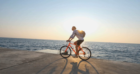 Fototapeta premium Young handsome male in casual wear ride on the colorful bicycle on the morning beach against beautiful sunset and the sea