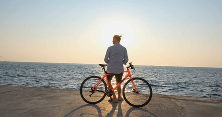 Young handsome male in casual wear ride on the colorful bicycle on the morning beach against beautiful sunset and the sea