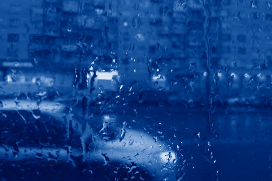 Misted glass with drops of rain. Behind the glass, the lights of the evening city. Painted in the classic blue color trend 2020. Blurry focus.