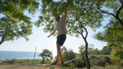 Athlete walking in slackline in the park with sea and blue sky on background	