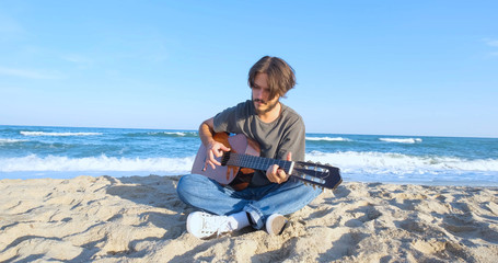 Young handsome male play in acoustic guitar on the beach in sunny day, sea or ocean on background 