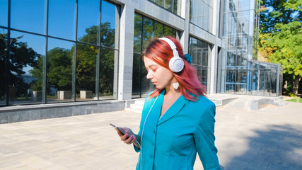 Young happy woman with pink hair and colorful suit walk outdoors and listen to music with headphones	