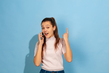 Pointing up with smartphone, smiling. Caucasian teen girl's portrait on blue background. Beautiful model in casual wear. Concept of human emotions, facial expression, sales, ad. Copyspace. Looks happy