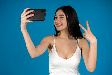 Smiling brunette girl taking selfie with peace sign isolated on blue background