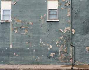 An exterior building wall painted with peeling gray green paint. Two windows and a downspout are also visible.