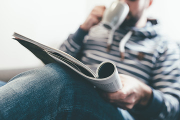 man reading magazine or newspaper and drinking coffee while relaxing on couch - Powered by Adobe