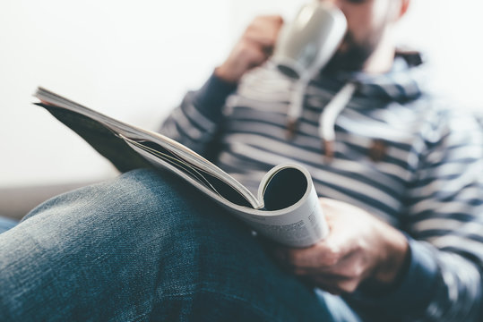 man reading magazine or newspaper and drinking coffee while relaxing on couch