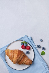 Fresh  croissant on a white plate and a blue napkin with berries  on a blue background.
