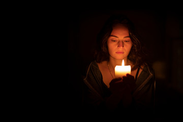 A sad religious orthodox brunette girl with brown eyes looks piercing and sad looking, holding a bright, lighted candle in folded hands. The girl on the right. Horizontal view