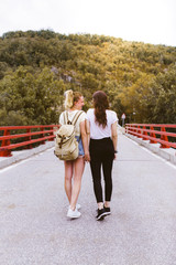 Back view of young girlfriends in love with backpack on the road near the mountain. Couple holding...