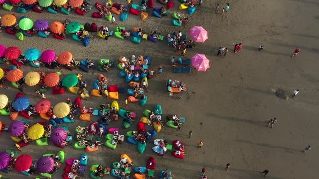 tourists relaxing on the multicolored beanbags under colorful umbrellas on the tropical sandy beach. Aerial high angle shot, double six beach, Kuta, Bali Indonesia
