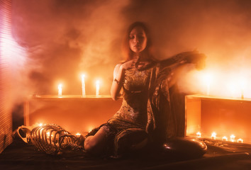 a witch with a snake on her neck and in her hands sits in a haze against a background of candles...
