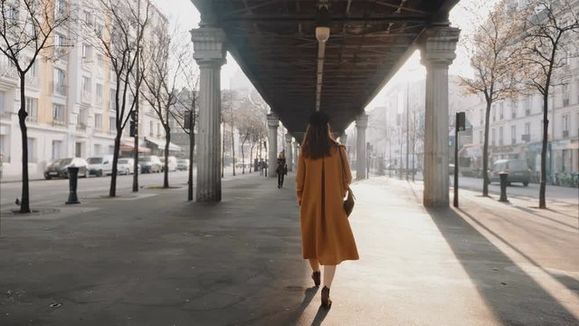 Wide shot camera follows stylish beautiful woman walking along Paris street under high line metro overpass slow motion.
