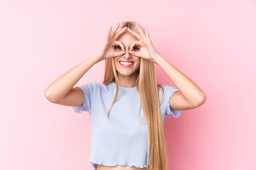 Young blonde woman on pink background showing okay sign over eyes