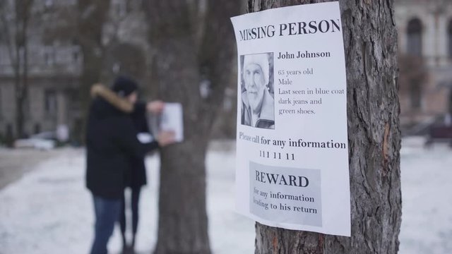 Close-up of missing person ad with information about lost senior Caucasian man. Blurred people hanging ads on trees at the background. Family searching for their grandfather. Unity, loss, kidnapping.
