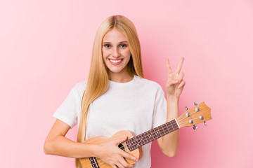 Young blonde girl playing ukelele showing victory sign and smiling broadly.