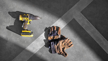 Sunlight and shadow on surface of the old yellow electric drill with brown leather gloves and white striped on concrete floor at workshop, Top view with copy space