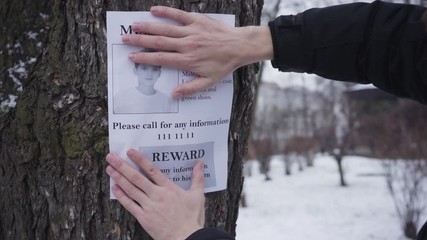 Bearded Caucasian man hanging missing boy ad on the tree. Young father searching for his lost son. Kidnapping, despair, social problems.
