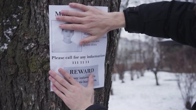 Bearded Caucasian man hanging missing boy ad on the tree. Young father searching for his lost son. Kidnapping, despair, social problems.