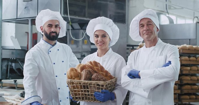 Smiling in front of the camera three bakers in a uniform in a big bakery industry holding a vintage basket with organic bread