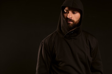 Model tests of a man on a black background, wearing a tracksuit. The gaze is lowered to the bottom of the frame. A beautiful beard and mustache on the face, and the head is covered with a hood