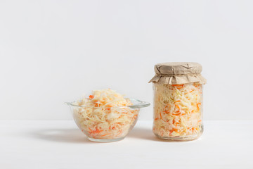 Homemade sauerkraut. Fermented food. Sauerkraut with carrots in glass jar and bowl with fork on wooden white background