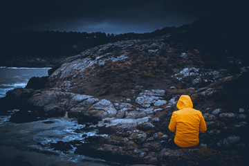 A young man adventurer back shot with contrast yellow jacket sitting over a rock by high cliff with...