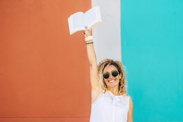 Read and reading paper book to study or relax in leisure activity concept with adult beautiful caucasian blonde curly woman giving up the book - coloured urban background and happy people