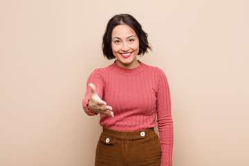 young pretty woman smiling, looking happy, confident and friendly, offering a handshake to close a deal, cooperating against beige wall
