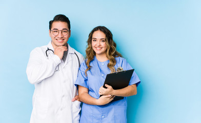 Young doctor couple posing in a blue background isolated smiling happy and confident, touching chin with hand.