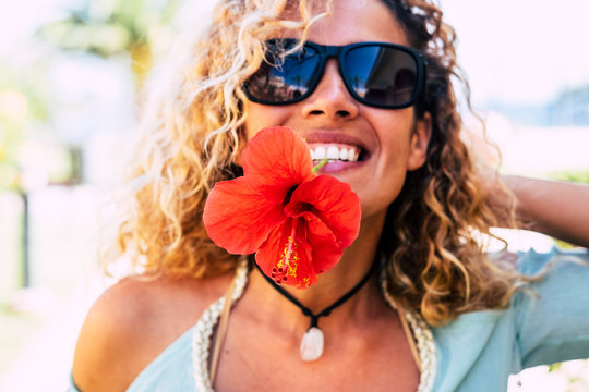 Spring and happiness concept with happy cheerful lady taking a big red flower with teeth in the mouth smiling and playing in front of the camera - beauty and freshness with adult people outdoor