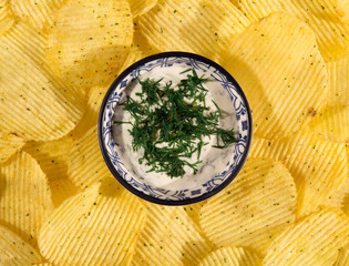 Potato chips on the table and in the center of a bowl with a blue ornament with sour cream and dill. Concept of unhealthy food.