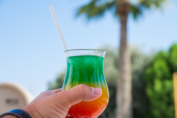 Vacation and rest concept. A man holds in his hand glass with colorful cocktail with drinking straw over the palm trees. Selective focus.