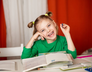 Naklejka premium little cute cheerful emotional girl, 7-8 years old, first grader, left-hander does homework at the table, at her desk, a lot of textbooks, blonde