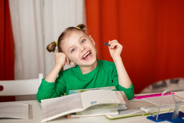 little cute cheerful emotional girl, 7-8 years old, first grader, left-hander does homework at the table, at her desk, a lot of textbooks, blonde