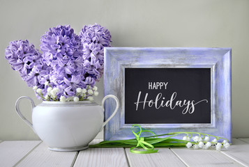 Blue springtime background. Light purple hyacinth and lily of the valley flowers around blackboard in violet frame. Text "Happy Holidays" on chalk board.