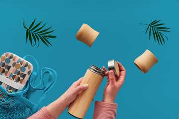 Zero waste tea in in eco friendly insulated bamboo flask. Two hands with cup and palm leaf, net bag with lunch box and tea flask are flying.Zero waste concept levitation on cyan, blue mint background.