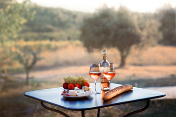 Bottle of rose wine and two full glasses of wine on table in heart of Provence, France with french bread, cheese, ham, grapes and peaches with olive trees on background - Powered by Adobe