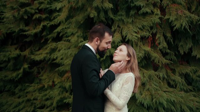 Groom hugs his bride, strokes her neck, head, hair, smiles at her touches her forehead against background of beautiful Christmas trees