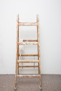 Indoor renovation scene. Wooden stepladder with paint splatters and hanging wipe on ladder rungs in front of white wall on carpet